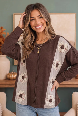 Woman wearing a cocoa brown knit top with taupe colorblock panels and vintage floral patches, styled with light wash jeans