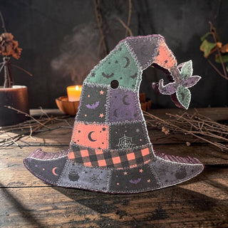A handmade felt witch hat decoration with a patchwork design of green, purple, orange, and dark gray sections, featuring embroidered moon, spiderwebs, and cauldron motifs, displayed on a rustic wooden surface with a warm, atmospheric background.