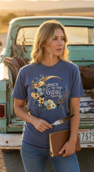 Woman wearing a blue t-shirt with floral design and text,  Text reads back to the Gyspsy that I Was standing in front of a vintage truck.