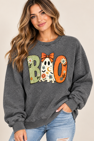A woman wearing a dark gray crewneck sweatshirt with a 'BOO' graphic made of Halloween-themed letters (a flowered 'B', a ghost 'I', and an orange 'O') paired with light blue jeans, set against a plain beige background.