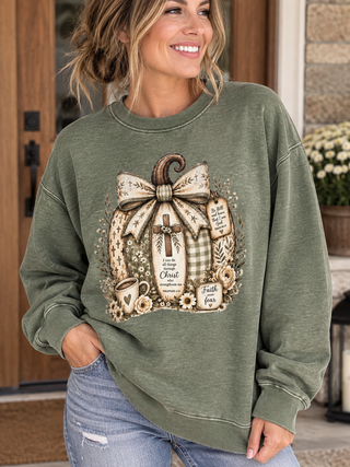 A woman wearing an olive green crewneck sweatshirt with a large autumn-themed graphic print featuring a pumpkin wreath, bow, cross, and text, paired with light blue jeans in front of a porch door.