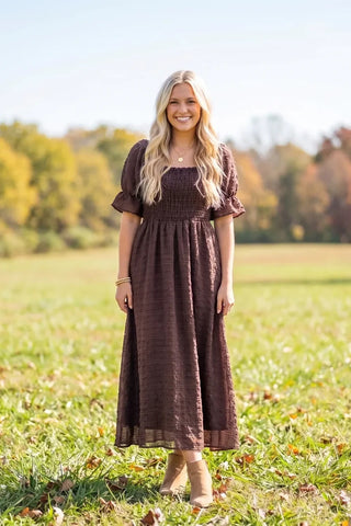 Model outdoors wearing a dark brown Simply Southern smocked maxi dress with puff sleeves and ankle boots.