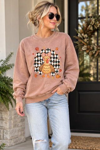 A woman wearing a dusty rose long-sleeve crewneck sweatshirt with a Thanksgiving turkey graphic, paired with light-wash distressed jeans and white sneakers, walking on a concrete porch.