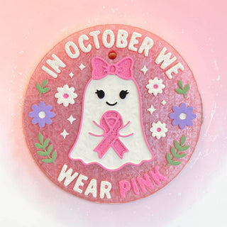 A round pink and white decorative ornament featuring a cute white ghost with a pink bow and pink awareness ribbon, surrounded by flowers and the text 'IN OCTOBER WE WEAR PINK'.